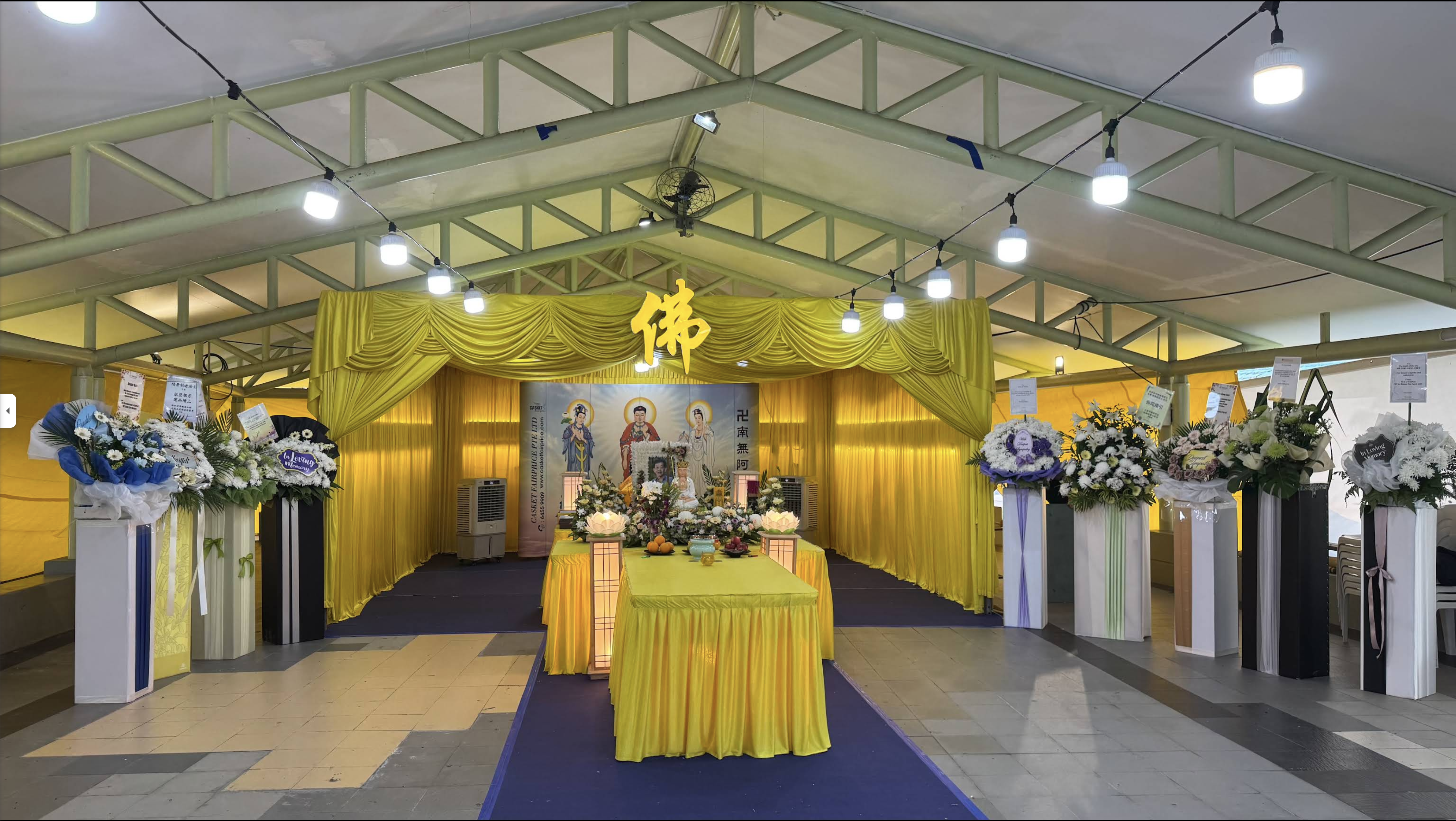 Funeral wake setup in a covered hall with an altar, floral tributes and offerings